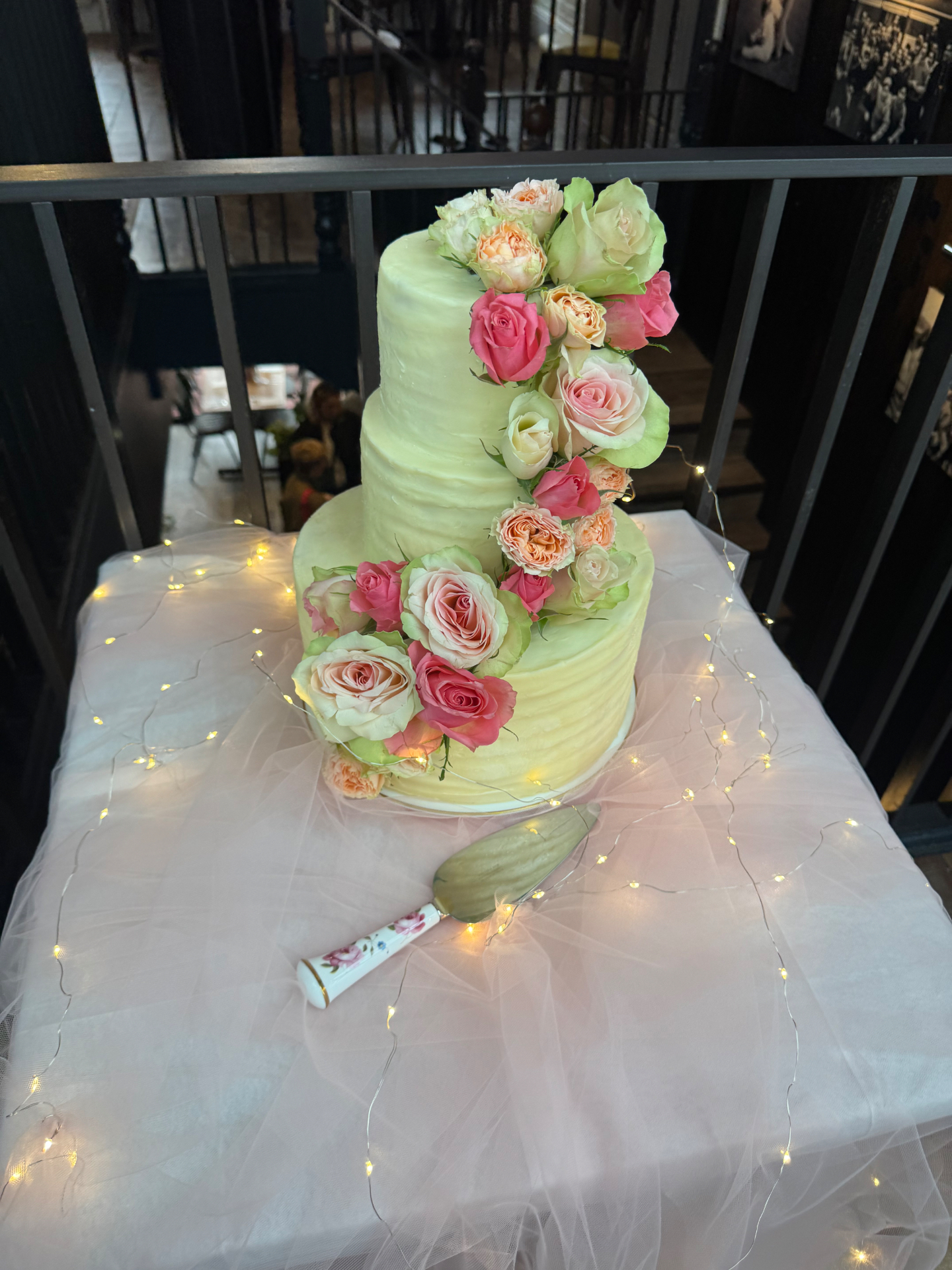 Three-tier pale yellow cake with cascading pink and peach roses and fairy lights
